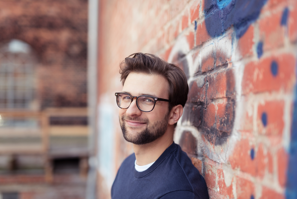 Portrait of Smiling Young Man with Facial Hair Wearing Eyeglasses and Leaning Against Brick Wall Painted with Graffiti
